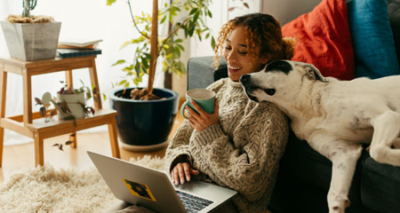 Young african american woman sitting on ground with dog on couch looking at laptop